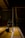 An indoor sports hall with a badminton net stand prominently positioned in the center. The scene is dimly lit, creating a dramatic contrast between the bright wooden floor and the dark background. A lone shuttlecock carrier with wheels is visible near the base of the net post.