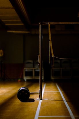An indoor sports hall with a badminton net stand prominently positioned in the center. The scene is dimly lit, creating a dramatic contrast between the bright wooden floor and the dark background. A lone shuttlecock carrier with wheels is visible near the base of the net post.