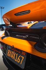 An orange sports car with a visible rear spoiler and McLaren branding. The image focuses on the back of the vehicle, showcasing a California license plate with the text '8XQX167'. The background includes a clear blue sky and portions of other parked cars.