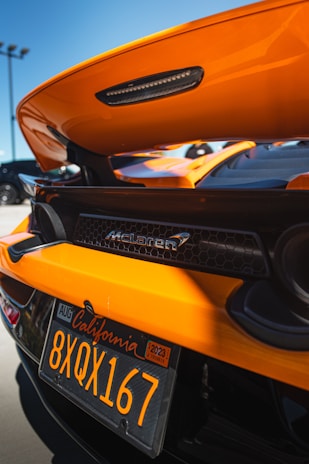 An orange sports car with a visible rear spoiler and McLaren branding. The image focuses on the back of the vehicle, showcasing a California license plate with the text '8XQX167'. The background includes a clear blue sky and portions of other parked cars.