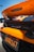 An orange sports car with a visible rear spoiler and McLaren branding. The image focuses on the back of the vehicle, showcasing a California license plate with the text '8XQX167'. The background includes a clear blue sky and portions of other parked cars.