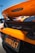 An orange sports car with a visible rear spoiler and McLaren branding. The image focuses on the back of the vehicle, showcasing a California license plate with the text '8XQX167'. The background includes a clear blue sky and portions of other parked cars.
