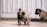A small brown puppy is walking on a tiled floor with curiosity. Another similar puppy is sitting in the background next to a green mat. The setting seems to be indoors with light-colored wooden walls.