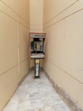 An old payphone is situated in a narrow, tiled hallway with beige walls. The phone booth is marked with 'World Call' branding and appears weathered, with visible wear on both the booth and the surrounding area.
