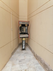 An old payphone is situated in a narrow, tiled hallway with beige walls. The phone booth is marked with 'World Call' branding and appears weathered, with visible wear on both the booth and the surrounding area.