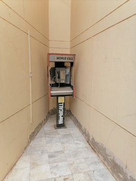 An old payphone is situated in a narrow, tiled hallway with beige walls. The phone booth is marked with 'World Call' branding and appears weathered, with visible wear on both the booth and the surrounding area.