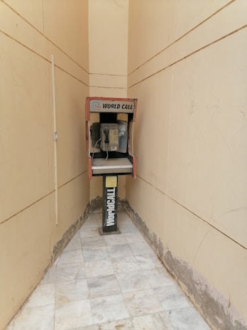 An old payphone is situated in a narrow, tiled hallway with beige walls. The phone booth is marked with 'World Call' branding and appears weathered, with visible wear on both the booth and the surrounding area.