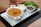 A steaming bowl of orange-colored soup garnished with herbs is placed on a white rectangular plate. Beside it are two small bowls, one filled with a light creamy sauce and the other with diced onions and herbs. Fresh green lettuce is also present on the plate.