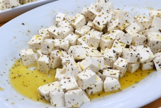 Cubed pieces of feta cheese are seasoned with herbs and drizzled with olive oil on a white plate.