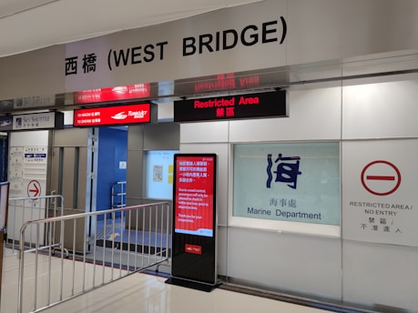 An airport or ferry terminal area with signage indicating a restricted area. There are signs in both English and Chinese stating 'Restricted Area' and 'Marine Department'. A digital display provides information about routes to Macau and Zhu Hai, alongside a warning about restricted entry and a visible crowd control barrier.