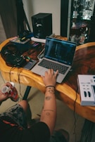 A person with tattoos on their arm works on a laptop placed on a wooden table. Nearby are various electronic devices including a speaker, headphones, and a MIDI controller. The setup is cluttered with wires and gadgets while the person holds a drink bottle. In the background, there's a glass cabinet with wine glasses.