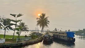 A tranquil waterway scene featuring a traditional houseboat docked near the shore during sunset. Palm trees line the shoreline, adding to the tropical ambiance. The sun is partially hidden behind clouds, casting a warm glow over the landscape.
