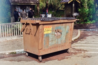 A large, rusted metal dumpster is placed in a parking area with clear signage indicating not to park in front of it. In the background, several people stand facing an information board with various notices and announcements surrounded by greenery and wooded areas.