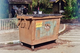 A large, rusted metal dumpster is placed in a parking area with clear signage indicating not to park in front of it. In the background, several people stand facing an information board with various notices and announcements surrounded by greenery and wooded areas.
