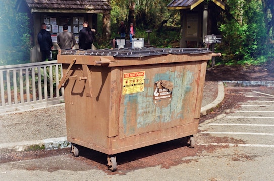 A large, rusted metal dumpster is placed in a parking area with clear signage indicating not to park in front of it. In the background, several people stand facing an information board with various notices and announcements surrounded by greenery and wooded areas.