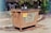 A large, rusted metal dumpster is placed in a parking area with clear signage indicating not to park in front of it. In the background, several people stand facing an information board with various notices and announcements surrounded by greenery and wooded areas.