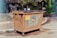 A large, rusted metal dumpster is placed in a parking area with clear signage indicating not to park in front of it. In the background, several people stand facing an information board with various notices and announcements surrounded by greenery and wooded areas.