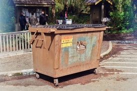 A large, rusted metal dumpster is placed in a parking area with clear signage indicating not to park in front of it. In the background, several people stand facing an information board with various notices and announcements surrounded by greenery and wooded areas.