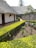 A serene garden with neatly trimmed green hedges, a variety of small plants and flowers, situated next to buildings with thatched roofs. The stone wall adds texture, and the path is clean and well-maintained. A fire extinguisher is mounted on one of the building walls.