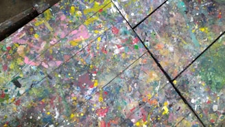 An abstract array of colorful paint splatters and drips covers a wooden surface, creating a chaotic and vibrant pattern. The paint layers overlap and blend, forming a rich tapestry of colors.