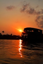 A serene sunset over water with a silhouette of a traditional houseboat. Palm trees line the distant shore, and the sky is painted with warm hues of orange and red.