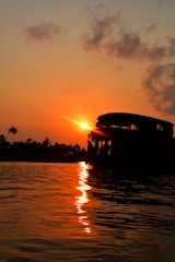 A serene sunset over water with a silhouette of a traditional houseboat. Palm trees line the distant shore, and the sky is painted with warm hues of orange and red.