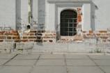 A weathered brick wall with patches of peeling paint and a section of old white plaster is accompanied by a metal-barred window. An aged downspout is attached to the wall, and the ground is covered with stone tiles with visible weeds growing from the cracks.