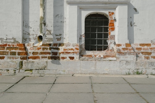 A weathered brick wall with patches of peeling paint and a section of old white plaster is accompanied by a metal-barred window. An aged downspout is attached to the wall, and the ground is covered with stone tiles with visible weeds growing from the cracks.