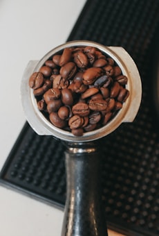 A close-up view of a portafilter filled with roasted coffee beans, placed on a textured black surface. The focus is on the rich, brown beans, highlighting their smooth, glossy surfaces and variation in color and texture.