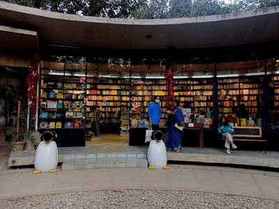 An outdoor bookstore with a curved concrete structure filled with colorful books on shelves. Two penguin statues stand at the entrance, and three people are present—one browsing books, another standing, and a third sitting on a bench. Red decorations hang from the ceiling.