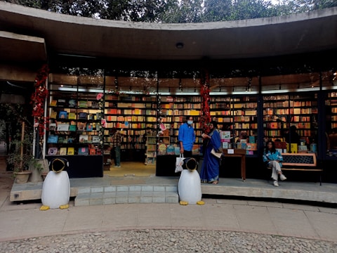 An outdoor bookstore with a curved concrete structure filled with colorful books on shelves. Two penguin statues stand at the entrance, and three people are present—one browsing books, another standing, and a third sitting on a bench. Red decorations hang from the ceiling.