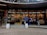 An outdoor bookstore with a curved concrete structure filled with colorful books on shelves. Two penguin statues stand at the entrance, and three people are present—one browsing books, another standing, and a third sitting on a bench. Red decorations hang from the ceiling.