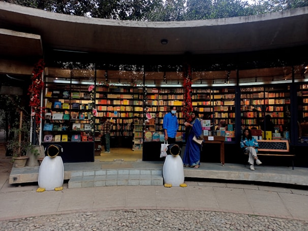 An outdoor bookstore with a curved concrete structure filled with colorful books on shelves. Two penguin statues stand at the entrance, and three people are present—one browsing books, another standing, and a third sitting on a bench. Red decorations hang from the ceiling.