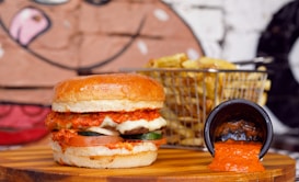 A juicy burger with layers of toppings such as melted cheese, sliced tomatoes, and cucumbers is placed on a wooden board. Beside it, there is a metal basket filled with golden-brown fries. A small black cup is overturned, spilling a bright red sauce onto the board. The background features a mural with a playful design.