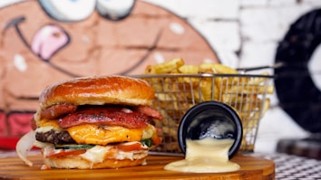 A juicy burger stacked with various layers including a cheese slice, tomato, meat patty, and pickles is placed on a wooden surface. Next to the burger is a wire basket filled with golden fries, with a small black dish slightly tipped to the side, spilling a creamy sauce. In the background, a mural featuring a cartoon face is partially visible.