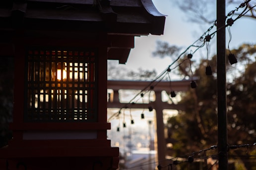 A traditional lantern with intricate lattice work is illuminated by the warm glow of the setting sun. Strings of small hanging lights create a festive atmosphere, while the silhouette of a torii gate appears in the background, suggesting a cultural or religious setting.