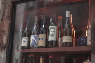 A collection of wine bottles displayed on wooden shelves. The bottles feature various unique and artistic labels, including illustrations of a human face and abstract designs. The setting appears rustic, with a textured glass and wooden framing partially obscuring the view.