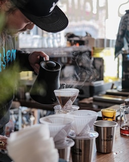 A person is carefully pouring hot water from a black kettle into a glass dripper filled with coffee grounds, as steam rises up. The setting appears to be a cozy, well-organized cafe or kitchen, with a counter displaying various coffee equipment, cups, and a small orange glass. The atmosphere is relaxed and focused, with soft lighting coming through a window.