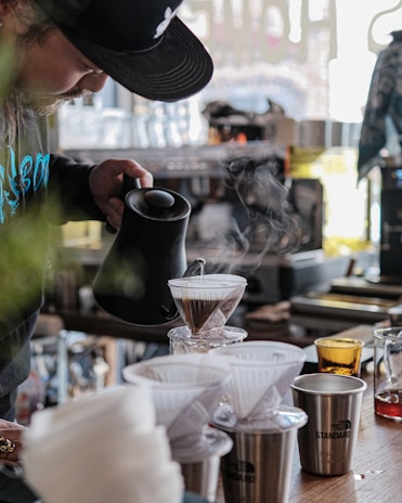 A person is carefully pouring hot water from a black kettle into a glass dripper filled with coffee grounds, as steam rises up. The setting appears to be a cozy, well-organized cafe or kitchen, with a counter displaying various coffee equipment, cups, and a small orange glass. The atmosphere is relaxed and focused, with soft lighting coming through a window.