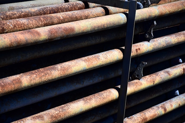 A close-up view of several rusted metal pipes stacked horizontally with dark brackets securing them. The pipes have a rough texture and display a mix of brown, orange, and greenish patina due to oxidation. Shadows and highlights create a textured, industrial look.