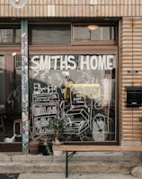 A storefront with a large window displays the words 'Smiths Home' and various white line drawings depicting household items and plants. Outside, there is a wooden bench and several potted plants. The building has a tiled facade with various stickers on the door frame.