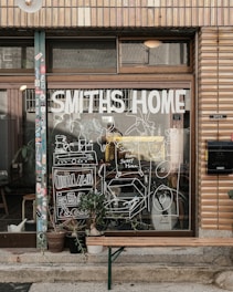 A storefront with a large window displays the words 'Smiths Home' and various white line drawings depicting household items and plants. Outside, there is a wooden bench and several potted plants. The building has a tiled facade with various stickers on the door frame.