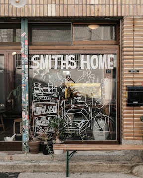 A storefront with a large window displays the words 'Smiths Home' and various white line drawings depicting household items and plants. Outside, there is a wooden bench and several potted plants. The building has a tiled facade with various stickers on the door frame.