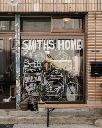 A storefront with a large window displays the words 'Smiths Home' and various white line drawings depicting household items and plants. Outside, there is a wooden bench and several potted plants. The building has a tiled facade with various stickers on the door frame.