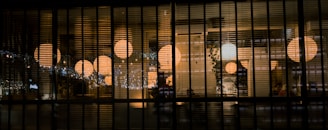 A dimly lit interior view through a series of vertical blinds, revealing warm glowing spherical lamps inside a room. The warm light contrasts with the dark tones of the blinds and exterior. Several people and decorative elements like plants and patterned walls are visible in the softly illuminated space.