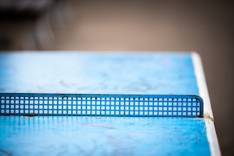 A close-up view of a blue table tennis table with a metal net in the center. The table surface has some wear and scratches, and the background is blurred, giving a shallow depth of field effect.