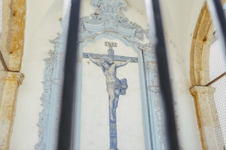 A tiled wall with a depiction of Christ on the cross is framed by ornate architectural features. The image is partially obscured by vertical metal bars in the foreground.