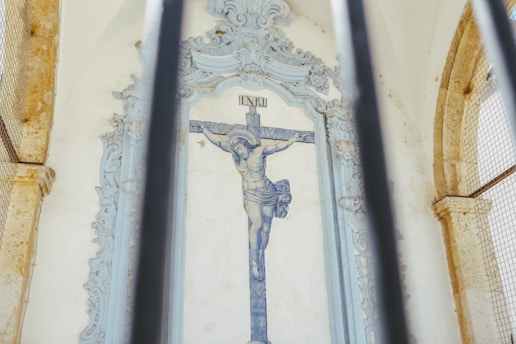 A tiled wall with a depiction of Christ on the cross is framed by ornate architectural features. The image is partially obscured by vertical metal bars in the foreground.