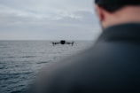 A drone is flying over an expansive body of water, with a person's shoulder visible in the foreground. The sky is overcast, suggesting a calm and contemplative setting.