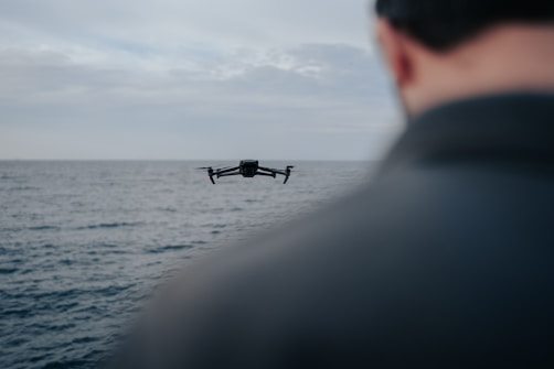 A drone is flying over an expansive body of water, with a person's shoulder visible in the foreground. The sky is overcast, suggesting a calm and contemplative setting.